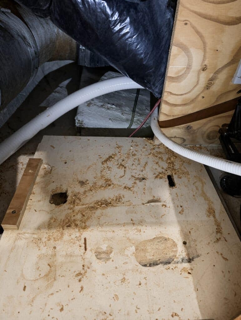 Plywood mounting board prepared for a Marine-Puck installation, with carriage-bolt holes, sawdust from fitting, and white flexible conduit run beside the bulkhead.